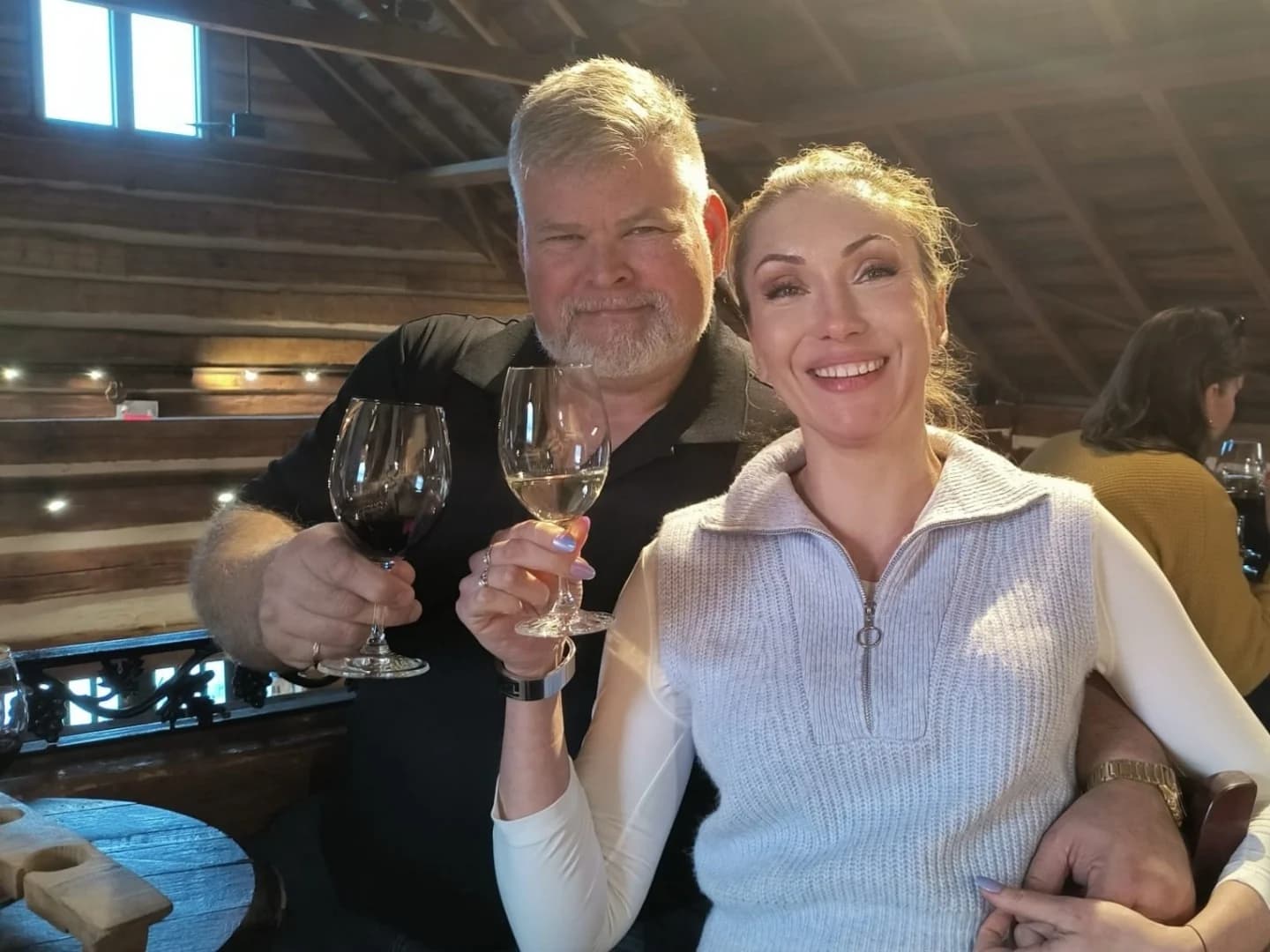 Robb and his girlfriend raising a glass of red and a glass of white at a table in a timber-framed Niagara winery tasting room